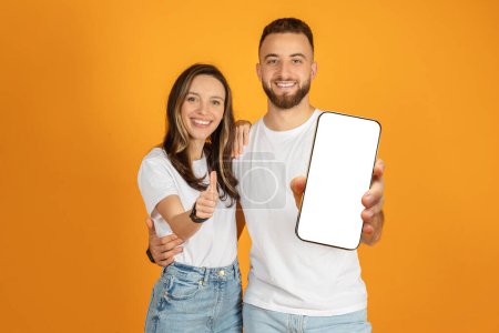 Téléchargez les photos : Un jeune couple européen joyeux donnant chacun un pouce levé, avec l'homme tenant un smartphone à écran blanc vers la caméra, sur un fond orange chaud, studio. App et appareil - en image libre de droit
