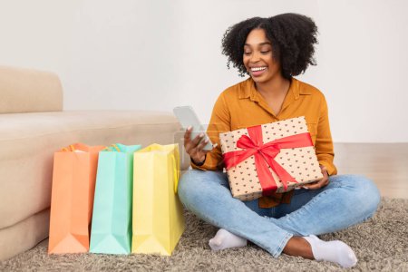Téléchargez les photos : Content heureuse femme afro-américaine millénaire tenant une boîte-cadeau enveloppée et regardant son smartphone, assis sur le sol à la maison avec des sacs à provisions colorés à côté d'elle - en image libre de droit