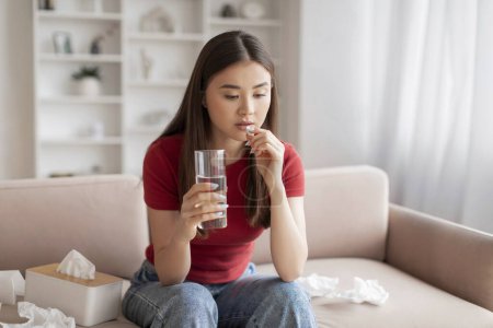 Foto de Preocupado joven mujer asiática tomando píldora con vaso de agua, señora coreana que sufre problemas de salud, sentado en el sofá rodeado de tejidos, que refleja el momento de autocuidado en casa, espacio de copia - Imagen libre de derechos