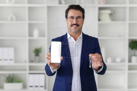 Foto de Hombre de negocios caucásico confiado en un traje azul marino sonriendo y presentando un teléfono inteligente con una pantalla en blanco y un conjunto de llaves, de pie en un espacio de oficina limpio y organizado - Imagen libre de derechos