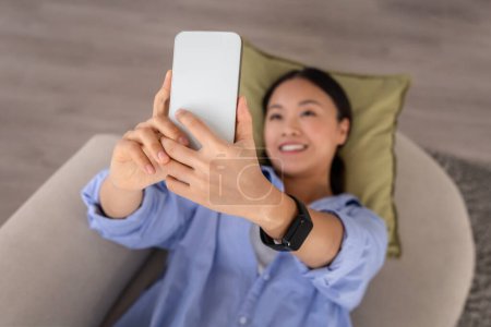 Téléchargez les photos : Belle femme chinoise millénaire prenant selfie sur un téléphone portable, couché sur le canapé à la maison, posant photo ou vidéo sur les médias sociaux, avoir une vidéoconférence avec des amis, vue sur le dessus - en image libre de droit