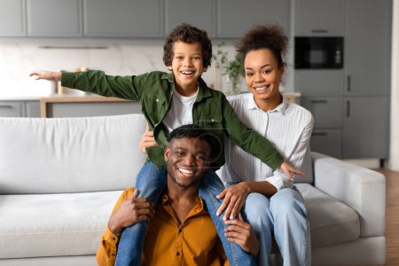 Photo for Cheerful young black boy sitting on his fathers shoulders, pretend fly, with mothers hand for support, embodying playful and loving family moment - Royalty Free Image