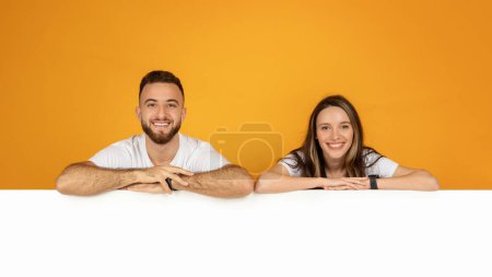 Téléchargez les photos : Un jeune homme et une jeune femme souriants européens s'appuient sur une surface horizontale blanche, grande bannière, à la recherche amicale et accessible, dans un contexte orange chaud, studio, panorama - en image libre de droit