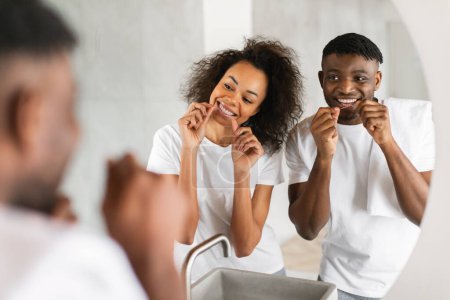 Téléchargez les photos : Afro-américain jeune couple utilise la soie dentaire, pratique les soins buccodentaires ensemble dans la salle de bain à la maison. Dentisterie familiale, hygiène quotidienne et concept de santé buccodentaire. Concentration sélective - en image libre de droit