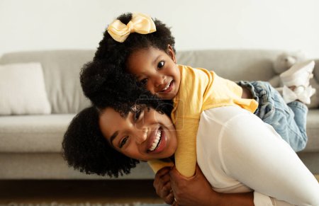 Téléchargez les photos : Mignon afro-américain petite fille tout-petit câlin et piggyback son heureux belle maman. joyeuse joyeuse mère noire et fille enfant s'amusent à la maison, embrasser et sourire à la caméra - en image libre de droit
