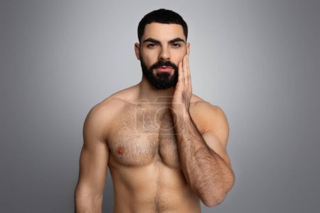Téléchargez les photos : Concept de routine de soins de la barbe. Attrayant millénial mec arabe seins nus touchant sa barbe, isolé sur fond gris. Moyen-Orient jeune homme jouissant de sa peau douce et soyeuse après les soins du visage - en image libre de droit