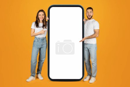 Téléchargez les photos : Un jeune homme et une jeune femme heureux d'Europe pointent vers un smartphone géant avec un écran vide, parfait pour l'application ou l'affichage Web, sur un fond orange vif, pleine longueur - en image libre de droit