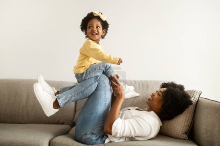 Photo for Side view of happy african american young mother and little girl playing together on couch at home interior, copy space. Mom enjoying time with her baby toddler. Motherhood concept - Royalty Free Image