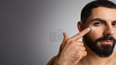 Photo for Men skin care concept. Closeup of handsome bearded middle eastern man putting face cream on his cheek, moisturizing skin after shower, isolated on grey studio background, panorama with copy space - Royalty Free Image