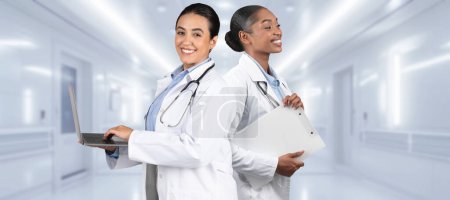 Téléchargez les photos : Deux joyeux professionnels de la santé afro-américains européens tiennent un ordinateur portable et un presse-papiers dans un couloir hospitalier lumineux, représentant les soins de santé modernes et le travail d'équipe - en image libre de droit