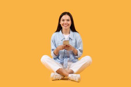 Téléchargez les photos : Joyeux jeune femme européenne en vêtements décontractés assis jambes croisées tenant smartphone, souriant à la caméra sur fond jaune vif - en image libre de droit