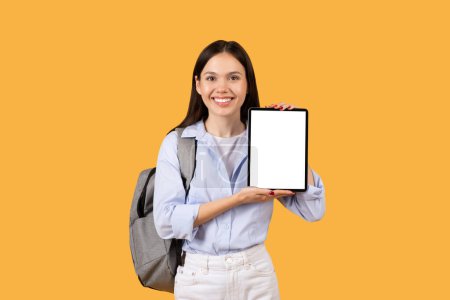 Photo for Smiling lady student in blue shirt holding tablet with white blank screen, showing educational app or website, against yellow background, ideal for copy space - Royalty Free Image