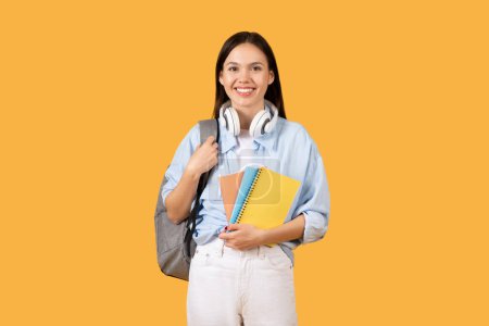 Téléchargez les photos : Étudiante rayonnante avec écouteurs et sac à dos gris portant des carnets colorés, posant sur fond jaune et souriant à la caméra - en image libre de droit