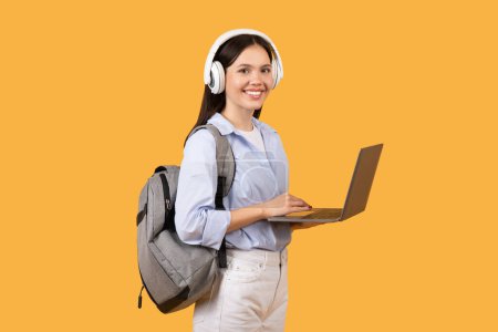 Photo for Cheerful female student using laptop while wearing white headphones and grey backpack, dressed in blue shirt, looking at camera against yellow background - Royalty Free Image