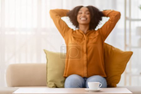 Téléchargez les photos : Une jeune femme afro-américaine exubérante aux cheveux bouclés s'étend les mains derrière la tête, souriant largement dans une pièce floue, avec une tasse blanche sur la table au premier plan - en image libre de droit