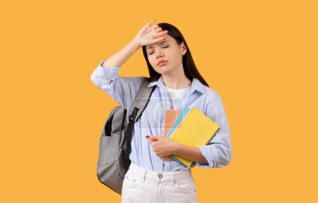 Photo for Weary female student touching forehead and holding notebooks, with grey backpack, tired from study, posing on yellow studio background - Royalty Free Image