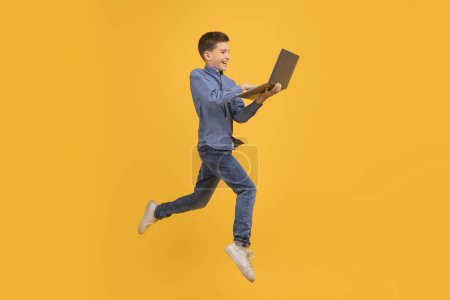 Téléchargez les photos : Image dynamique d'un adolescent sautant et tenant un ordinateur portable ouvert, adolescent homme utilisant un ordinateur tout en volant sur fond jaune, symbolisant l'agilité à l'ère numérique, bénéficiant d'une éducation en ligne - en image libre de droit