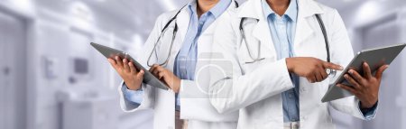 Téléchargez les photos : Deux professionnels de la santé en blouse blanche utilisent des tablettes numériques, probablement pour examiner les informations sur les patients, dans un cadre hospitalier moderne. Dispositif moderne pour les soins de santé - en image libre de droit