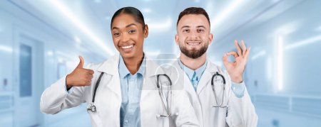 Téléchargez les photos : Deux joyeux professionnels de la santé caucasiens, afro-américains en blouse blanche donnent un pouce levé et signe correct, exsudant la confiance et la positivité dans un cadre hospitalier lumineux - en image libre de droit