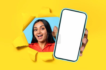 Photo for Excited indian young woman tears paper wall presenting mobile phone device, showing large empty screen recommending application through hole in yellow studio background. Collage, mockup - Royalty Free Image