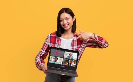 Foto de Una joven asiática sonriente con una camisa a cuadros roja apunta a la pantalla de su computadora portátil mostrando una amistosa reunión en línea con otros cuatro participantes sonrientes sobre un fondo amarillo - Imagen libre de derechos
