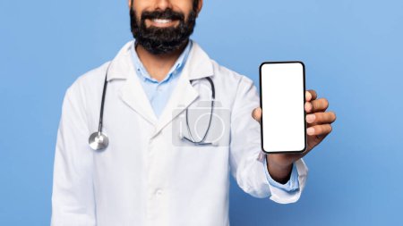 Foto de Sonriente médico indio con barba, mostrando un teléfono inteligente con pantalla en blanco, vistiendo bata blanca de laboratorio y estetoscopio, sobre fondo azul, publicidad médica - Imagen libre de derechos
