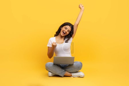 Téléchargez les photos : Joyeux émotionnel jolie jeune étudiante indienne avec ordinateur sur ses genoux assis sur le sol sur fond de studio jaune, levant la main et s'écriant, achète cours pas cher. Éducation en ligne - en image libre de droit