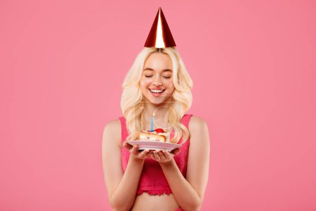 Photo for Joyous young blonde lady in party hat, cherishing moment with eyes closed, gently holding birthday cake with lit candle, against pink backdrop - Royalty Free Image
