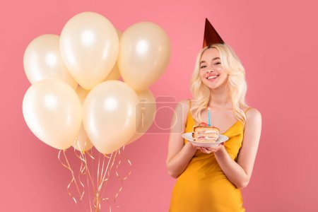 Foto de Mujer rubia alegre en sombrero de fiesta y vestido amarillo, celebración de pastel de cumpleaños con vela encendida, acompañado de un montón de globos de crema, fondo rosa - Imagen libre de derechos