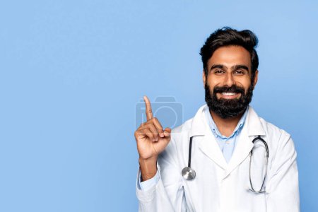 Photo for Happy middle aged indian male doctor with beard pointing his finger upwards, wearing ab coat and stethoscope, with cheerful expression on blue background - Royalty Free Image