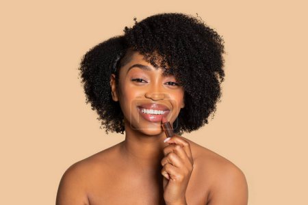 Téléchargez les photos : Joyeuse jeune femme afro-américaine avec des cheveux bouclés appliquant rouge à lèvres nu, regardant heureux avec un sourire lumineux, sur fond beige - en image libre de droit