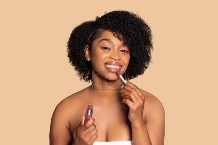 Foto de Alegre mujer afroamericana con el pelo rizado aplicando brillo labial rosa, sonriendo a la cámara, envuelta en toalla blanca contra fondo beige estudio - Imagen libre de derechos
