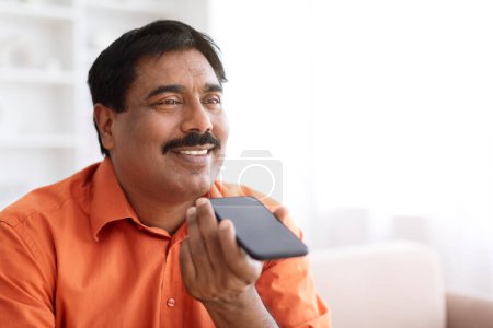 Téléchargez les photos : Joyeux homme indien mature avec moustache enregistrement de message vocal, en utilisant un smartphone à la maison, panorama avec espace de copie, en utilisant l'assistant AI pour la recherche sur le téléphone, gros plan - en image libre de droit