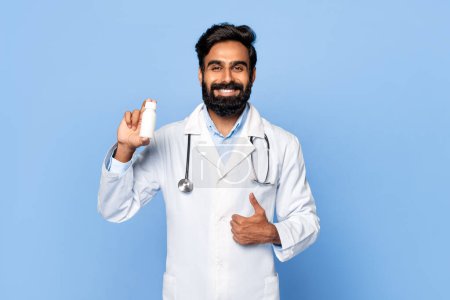 Photo for Cheerful indian male doctor with stethoscope around his neck, holding white pill bottle and giving thumbs-up sign, smiling at camera against blue background - Royalty Free Image