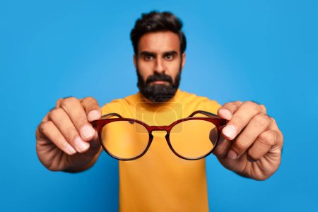 Photo for Serious middle aged indian man in yellow shirt holding out pair of glasses towards the camera, focus on spectacles against bright blue background - Royalty Free Image