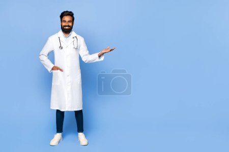 Téléchargez les photos : Médecin indien homme confiant avec stéthoscope autour du cou, gesticulant avec sa main pour montrer quelque chose d'invisible à l'espace libre, debout sur fond bleu - en image libre de droit