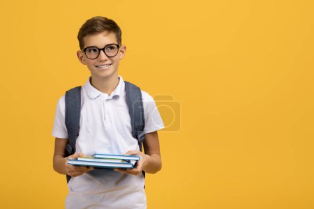 Photo for Intelligent schoolboy wearing glasses and backpack holding textbooks and looking aside at copy space, smiling male kid ready for school, standing against yellow studio background, panorama - Royalty Free Image