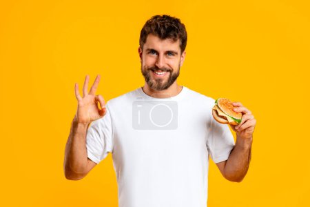 Photo for Junk food approval. Portrait of cheerful man in white t-shirt gesturing okay sign while holding takeaway burger, on yellow studio backdrop, smiling to camera. Customer feedback concept - Royalty Free Image