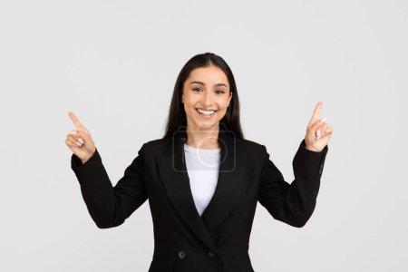 Radiant european businesswoman in tailored suit happily pointing upwards with both hands at free space, drawing attention to something important
