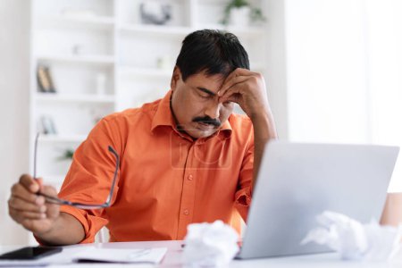 Foto de Exhausto hombre de negocios indio maduro que sufre de burnout, sentado en el escritorio frente a la computadora portátil, sosteniendo gafas, frotando su cabeza, interior de la oficina en casa. Bancarrota, crisis - Imagen libre de derechos