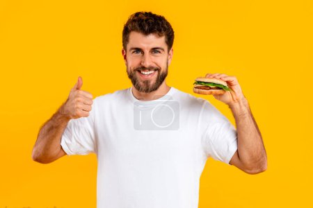 Photo for I like junk food. Portrait of cheerful bearded guy holding cheeseburger and gesturing thumbs up on yellow studio background, smiling to camera. Eater approving taste of favorite fastfood - Royalty Free Image