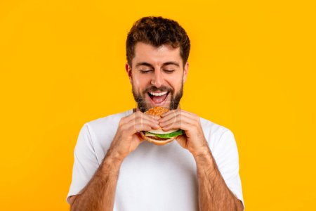 Photo for Cheat day concept with millennial guy enjoying tasty burger over yellow backdrop, ideal for nutrition advertising. Studio portrait of young caucasian man eating cheeseburger - Royalty Free Image