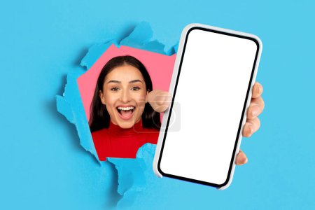 Photo for Excited caucasian young woman showing cell phone with big empty screen, offering mock up for mobile advertisement design, displaying smartphone through hole in blue paper backdrop. Collage - Royalty Free Image
