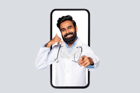 Téléchargez les photos : Médecin barbu masculin souriant affiché dans le cadre du smartphone faisant appel à moi geste, représentant les services de télémédecine, sur fond de lumière - en image libre de droit