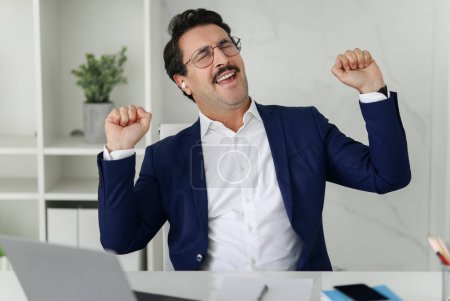 Téléchargez les photos : Homme d'affaires caucasien extatique en costume bleu avec les yeux fermés et les poings pompés célèbre une victoire ou un succès, ajoutant un sentiment de réussite et de joie à l'environnement de bureau - en image libre de droit