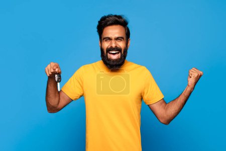 Photo for Overjoyed indian bearded man in yellow shirt holding up car keys and pumping his fist, expressing triumph and happiness, against blue background - Royalty Free Image
