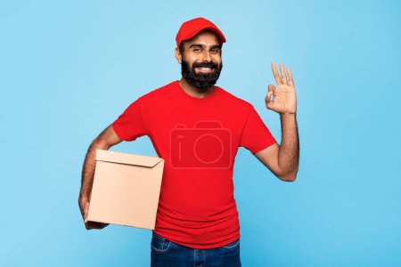 Foto de Repartidor indio sonriente con camisa roja y gorra que sostiene el paquete de cartón y hace un gesto aceptable, lo que significa un servicio confiable, contra el fondo azul - Imagen libre de derechos