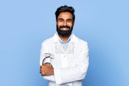 Photo for Cheerful bearded middle aged indian male doctor with crossed arms and stethoscope, wearing lab coat, radiating confidence against blue background - Royalty Free Image
