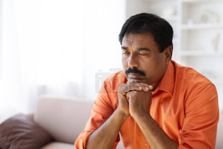Photo for Mental health. Closeup of sad mature indian man sitting on couch alone at home, leaning on his hands and looking down, experiencing difficulties in life, going through divorce, bankruptcy, copy space - Royalty Free Image