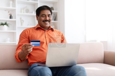 Photo for Mature indian man with moustache sitting on couch, using computer laptop and bank credit card at home, shopping or banking online, paying for goods and services, copy space. Easy banking, ecommerce - Royalty Free Image